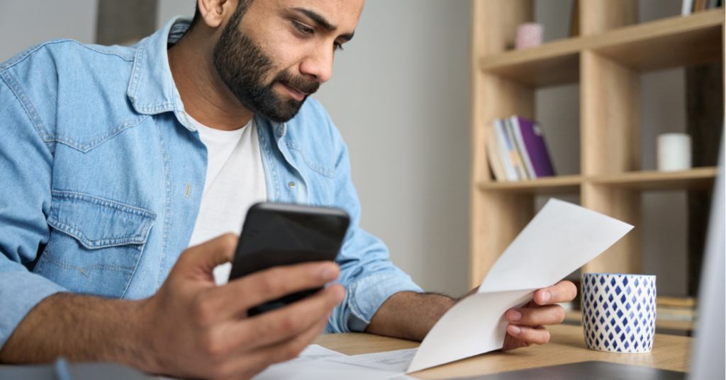 Critical Student Loan Repayment Changes for Dentists A man is reviewing student loan repayment changes for dentists on his phone while going through paper bills at his desk.