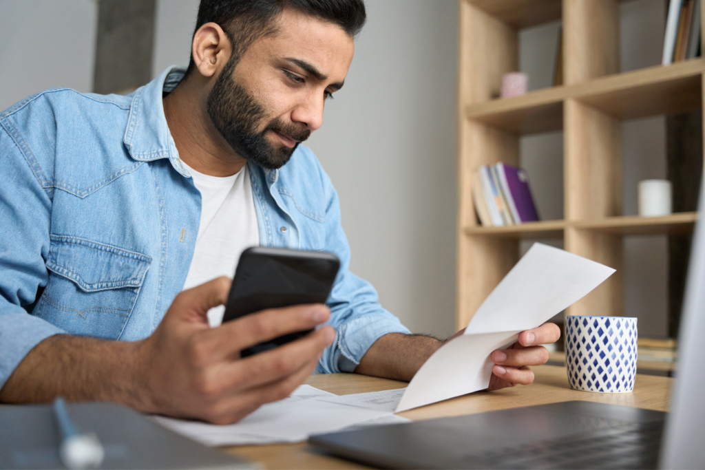 Critical Student Loan Repayment Changes for Dentists Dentist reviewing student loan repayment changes on his phone while going through paper bills at his desk.